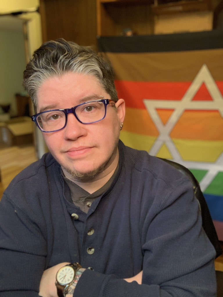 Photo of a white transgender man with short brown and gray hair, wearing glasses, sitting with arms crossed in front of a rainbow Pride flag with a Star of David on it